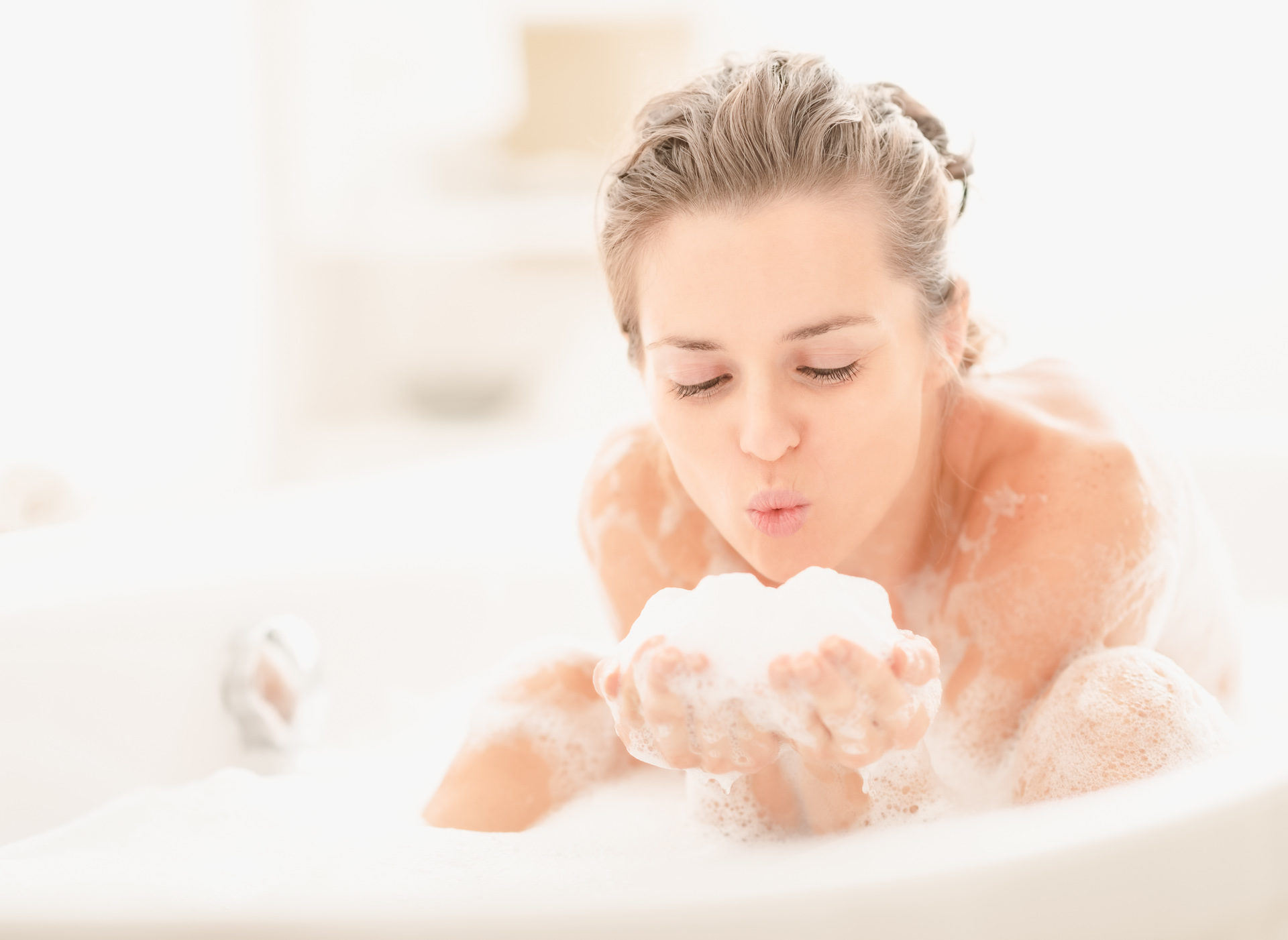 Eine Frau sitzt in der Badewanne und pustet auf Badeschaum in ihrer Hand.